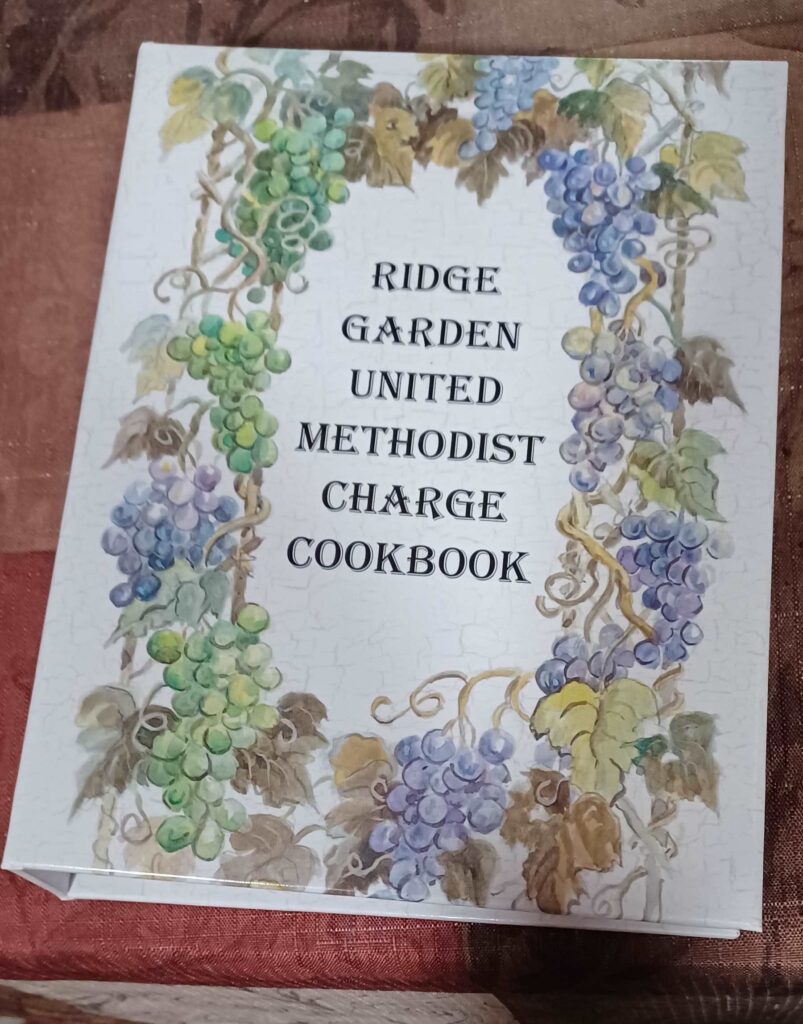 White book cover entitled: Clover Ridge Garden United Methodist Charge Cookbook with garland of grapes around it